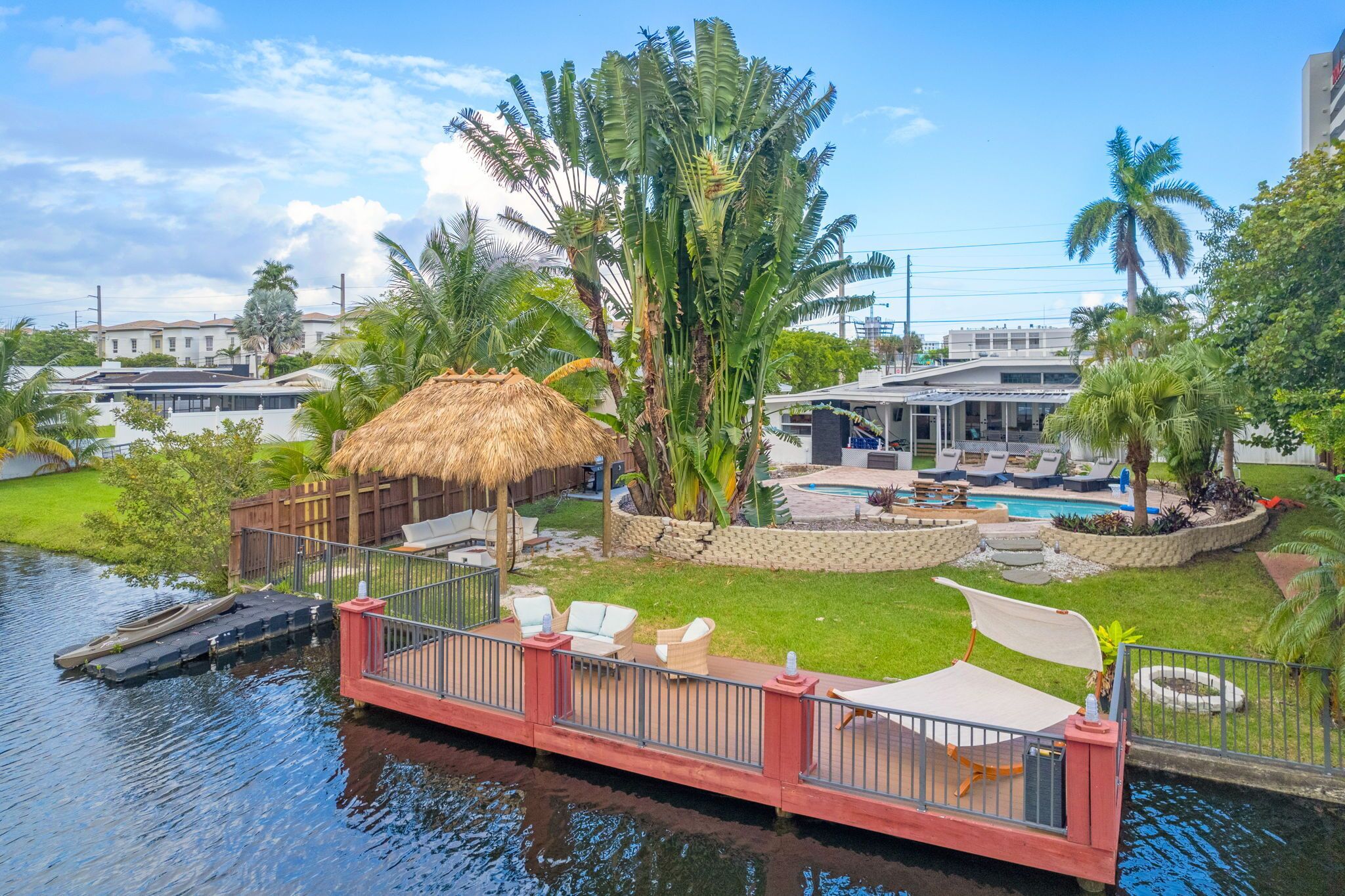 A waterfront property with a pool, lounge chairs, and a thatched-roof structure.