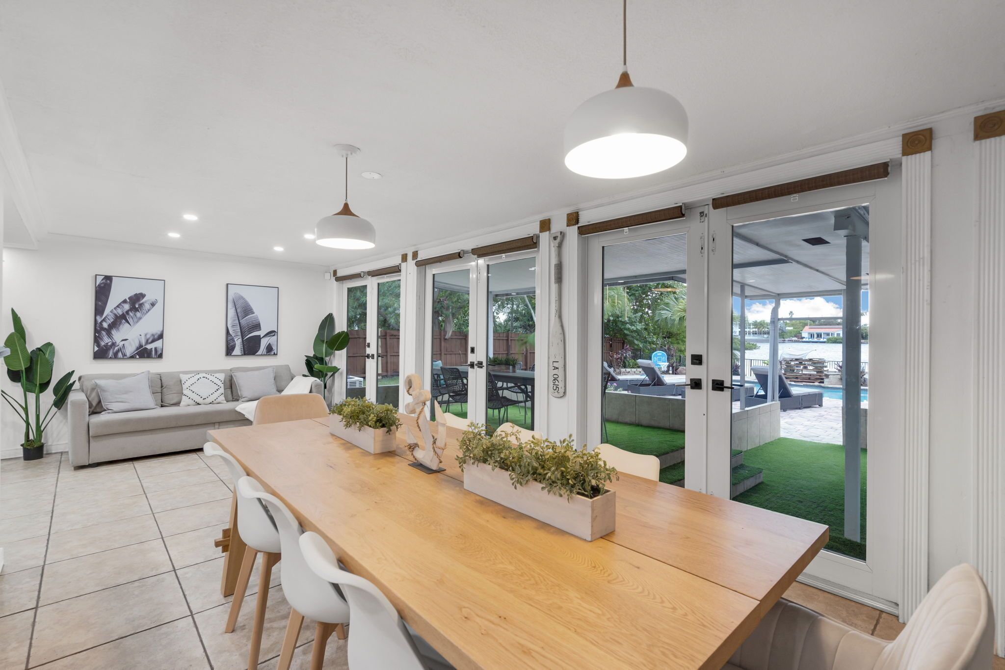 Um interior moderno com uma longa mesa de madeira, cadeiras brancas e um sofá. Há plantas em vasos sobre a mesa e vista para uma área externa com piscina.