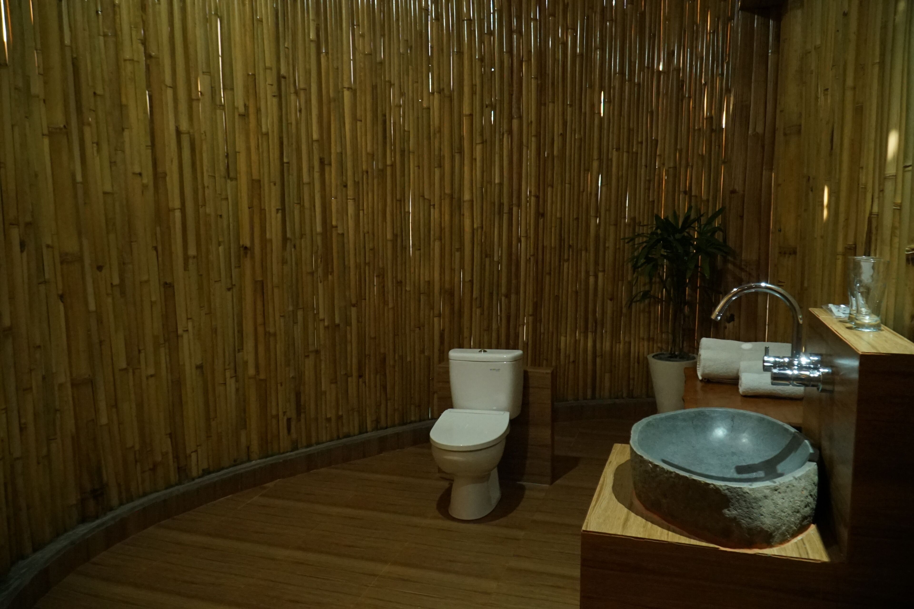 A bathroom with a bamboo wall, a toilet, a sink with a basin, and a potted plant.