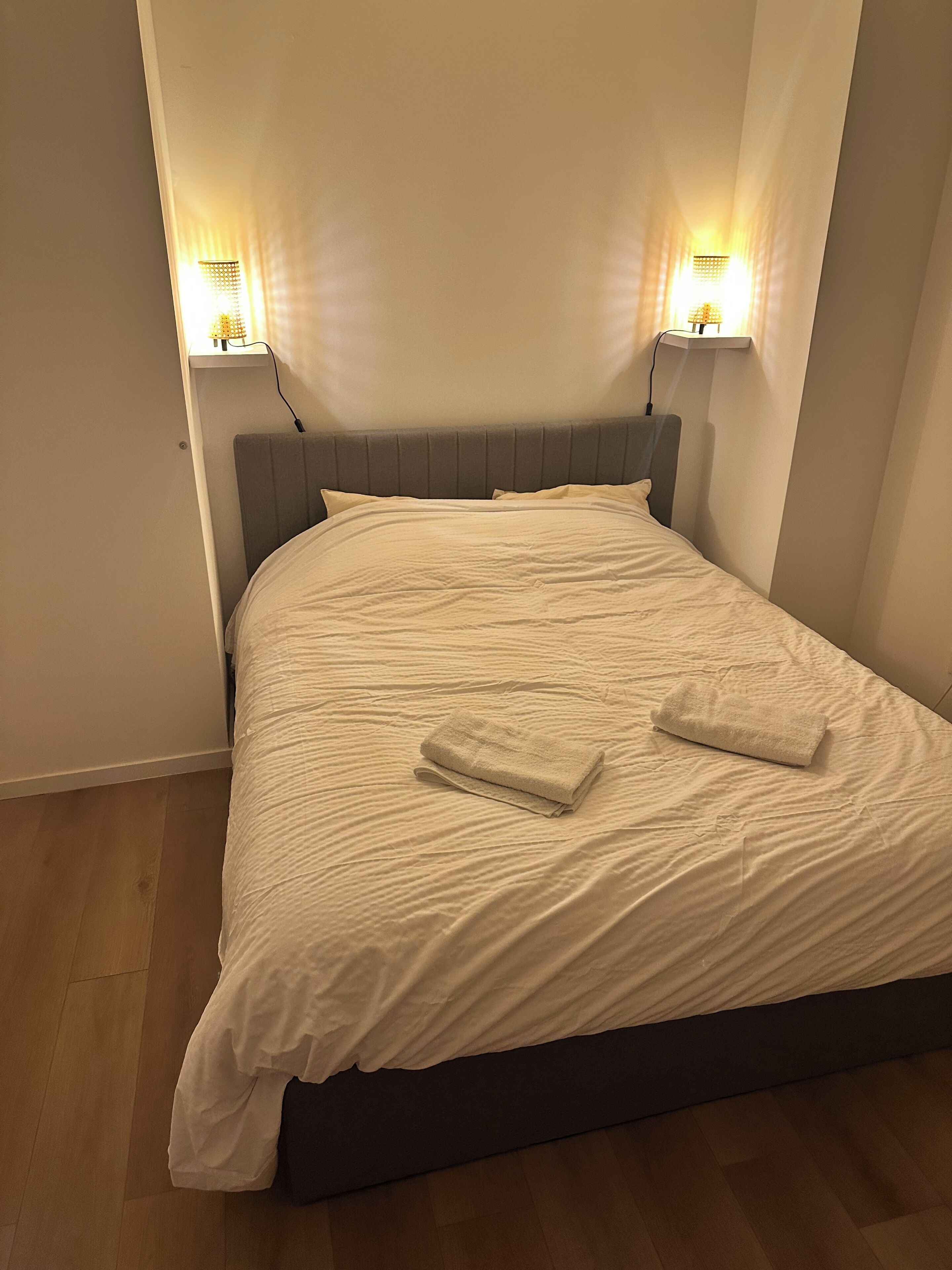 Una cama con cabecera, dos lámparas de noche y dos toallas dobladas.
