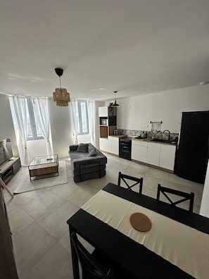 A modern living room with a sofa, a dining table, and a kitchen in the background. - T2 in the heart of Suze - Between Vineyards and History (Suze-la-Rousse)
