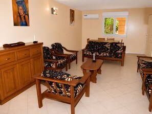 Ein Wohnzimmer mit einer Sofagarnitur, Holznachttischen und einem Schrank. Zwei Wandklimaanlagen und ein Fenster mit Blick auf Grünflächen.