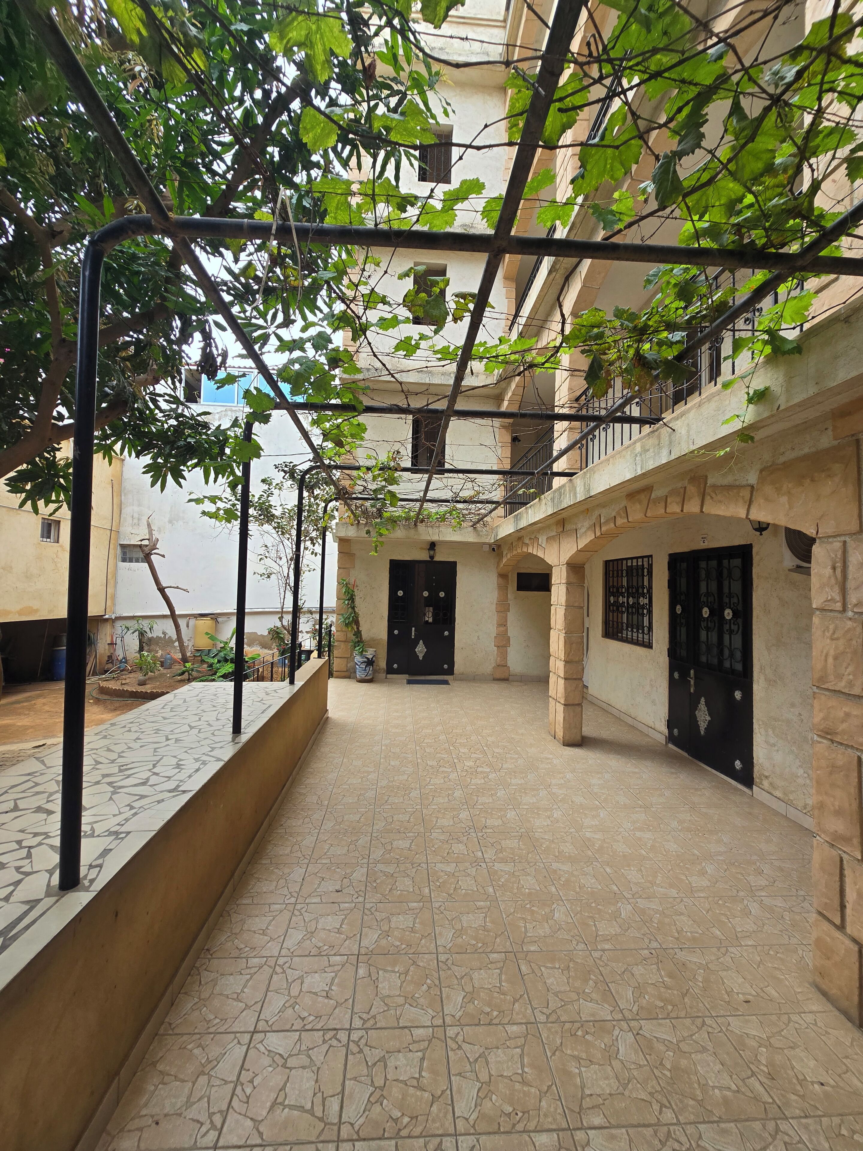 Un patio con suelo de baldosas, una pérgola de metal y un edificio con puerta negra y ventanas con barrotes.