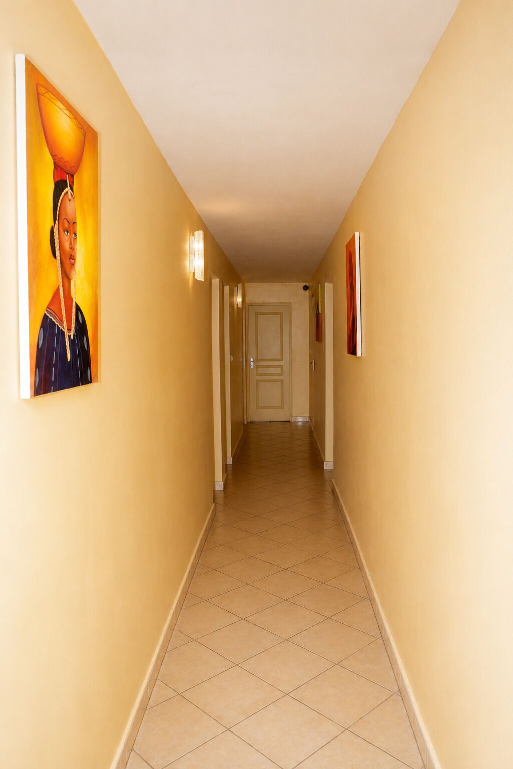A hotel corridor with a tiled floor, a door at the end, and wall-mounted light fixtures.