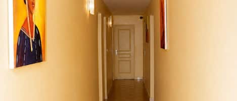 A hotel corridor with a tiled floor, a door at the end, and wall-mounted light fixtures.