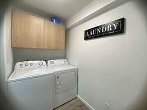 A laundry area with two white appliances, wooden cabinets, and a wall-mounted sign.