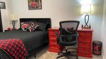 A bedroom with a bed, a desk, a chair, a lamp, a trash can, and a framed picture on the wall.