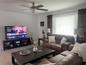 A living room with a flat-screen TV, a ceiling fan, a sofa, a coffee table, and a window with blinds.