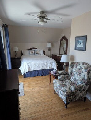 A hotel room with a bed, a chair, a desk, a lamp, a ceiling fan, and a picture on the wall.