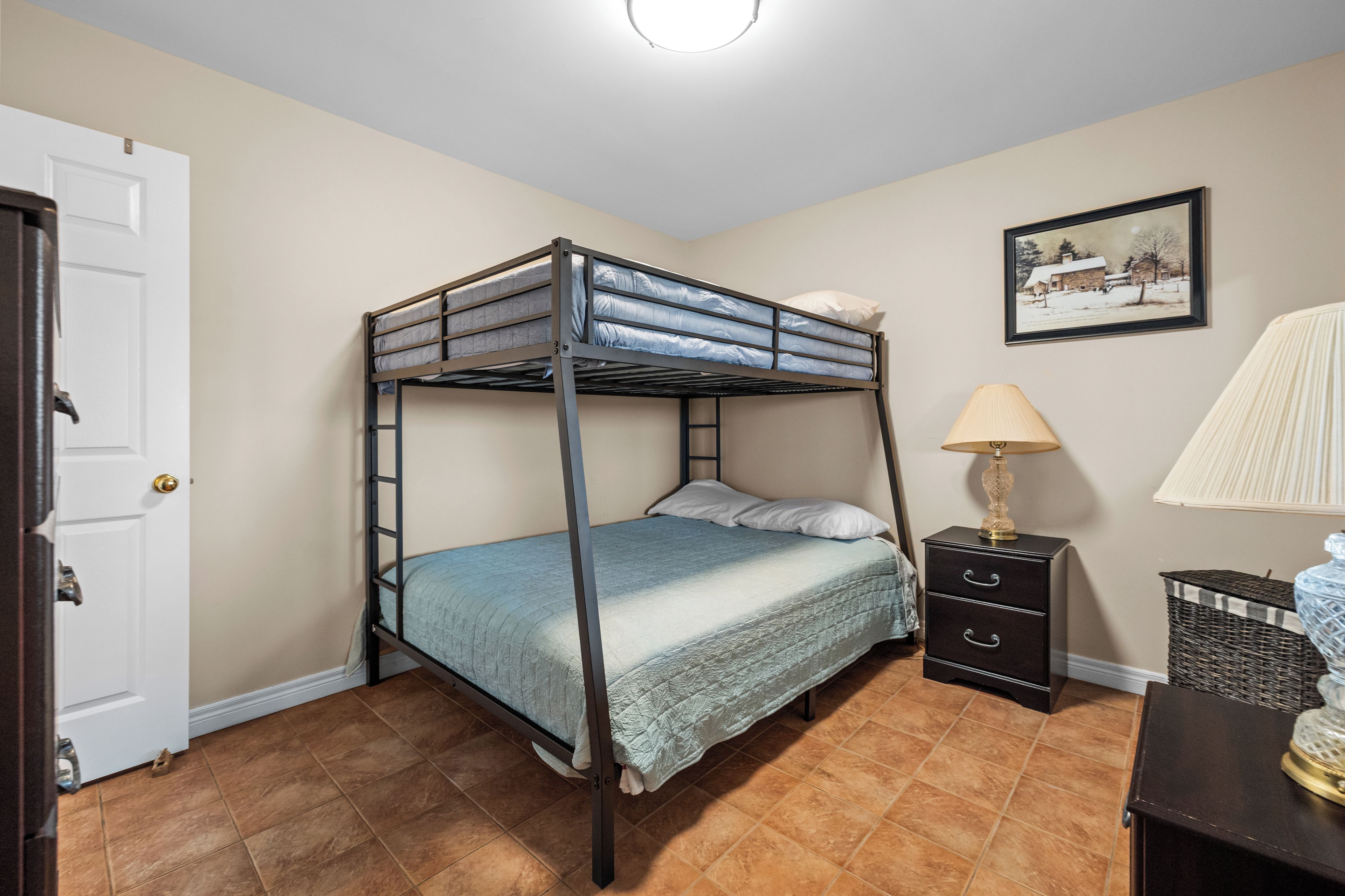 A bunk bed in a room with a door, a lamp, a picture on the wall, and a small table.