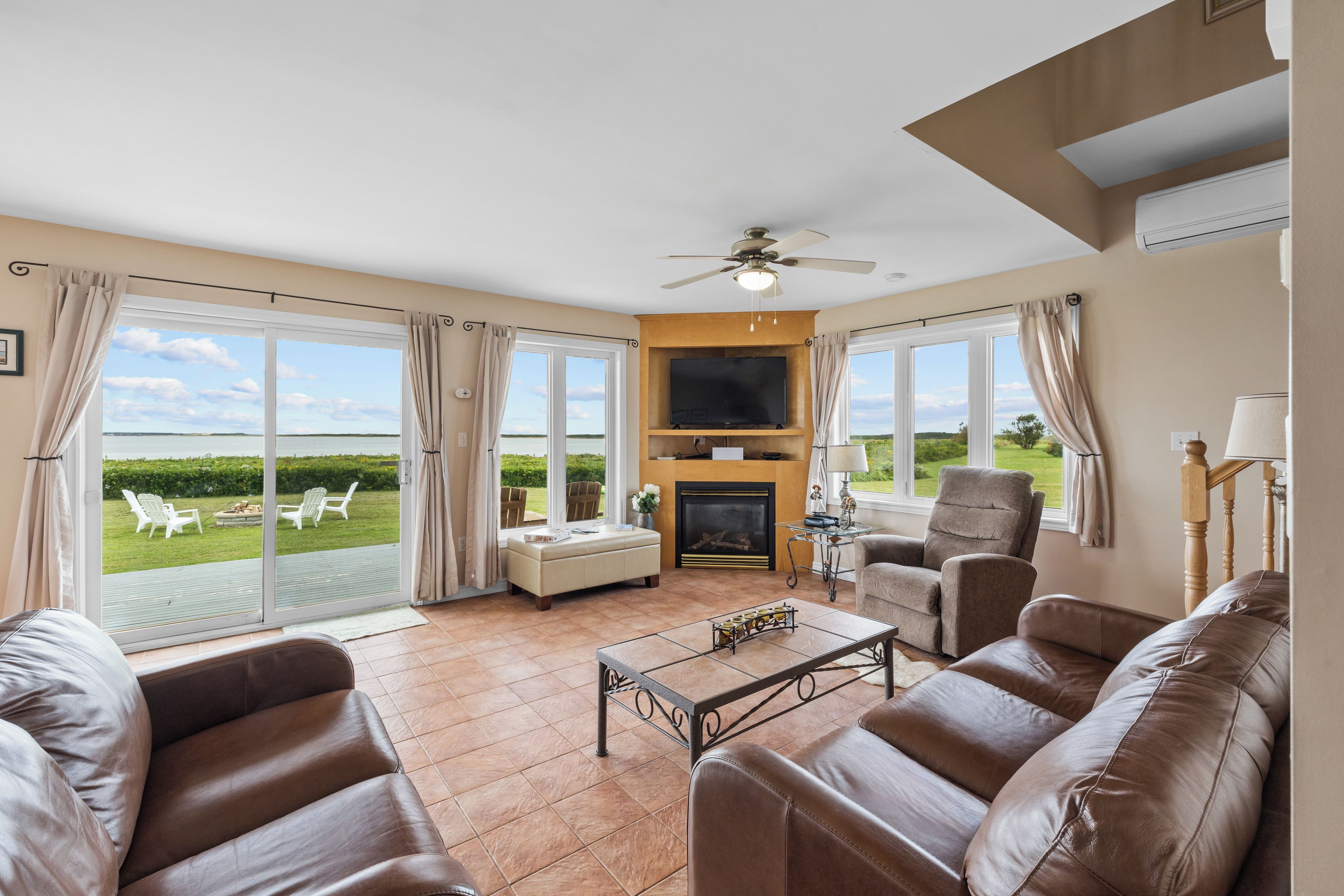 A spacious living room with leather sofas, a fireplace, a flat-screen TV, and large windows offering a view of the outdoors.