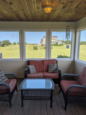 Une terrasse couverte, aménagée avec des meubles en osier, une table à plateau de verre, et offrant une vue sur une maison et un paysage verdoyant à travers de grandes fenêtres.