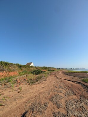 A coastal landscape with a dirt path leading to a white house on a hill, surrounded by vegetation and a clear sky.