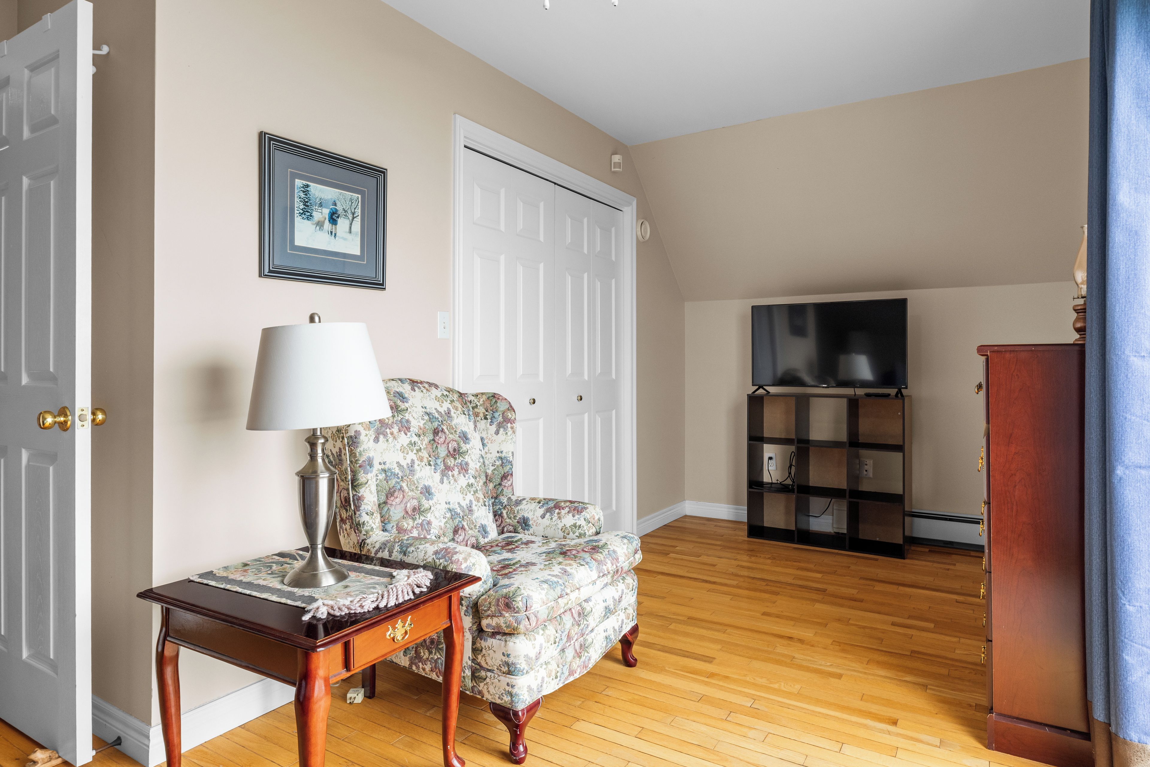 A living room with a floral armchair, a wooden coffee table, a television stand, and a wardrobe.
