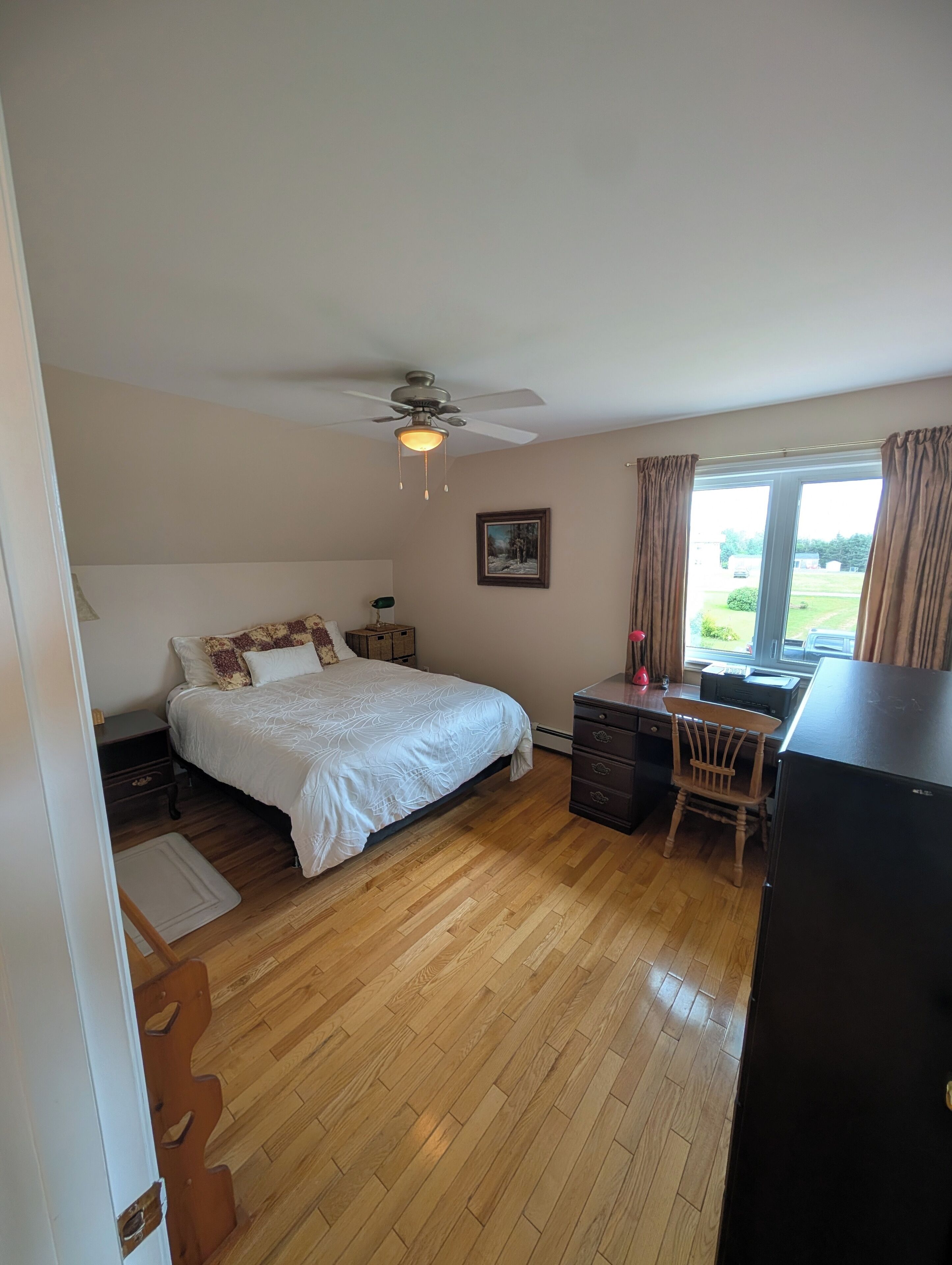 A bedroom with a bed, a desk, a chair, a ceiling fan, and a window.