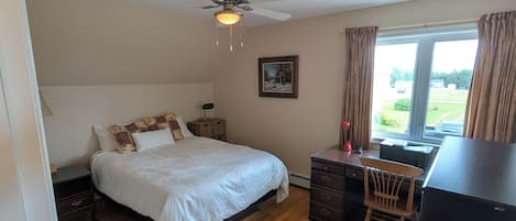 A bedroom with a bed, a desk, a chair, a ceiling fan, and a window.