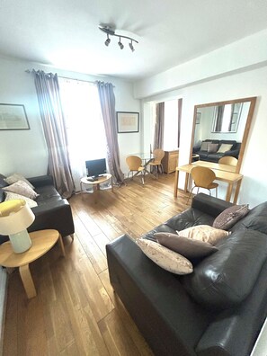 A furnished living room with a black leather sofa, a small round table, a television, and a dining area with chairs and a table. - Stylish Central London apartment is next to the tube Chelsea border (London)