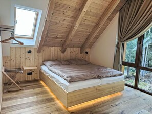 Ein Schlafzimmer mit schrägen Wänden, einem Bett mit Kopfteil, einem Fenster mit Vorhängen und einem Holzboden.