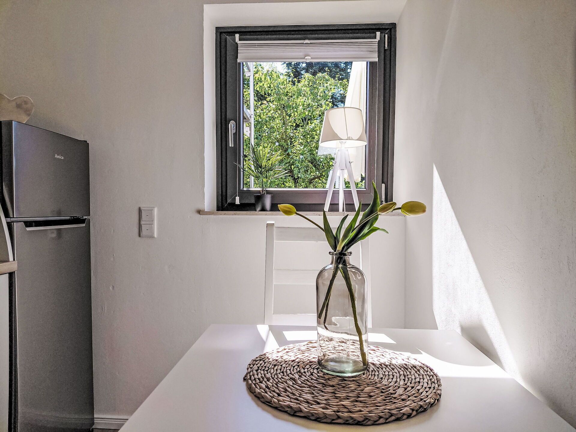 Cozinha compacta com geladeira, janela com vista para o verde e mesa com um vaso de flores.