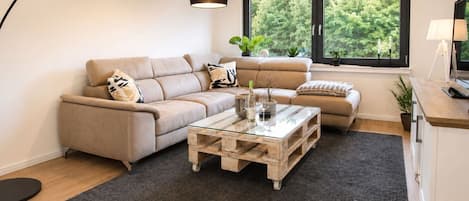 A modern living room with a sofa, a glass coffee table, and a view of greenery through a window.