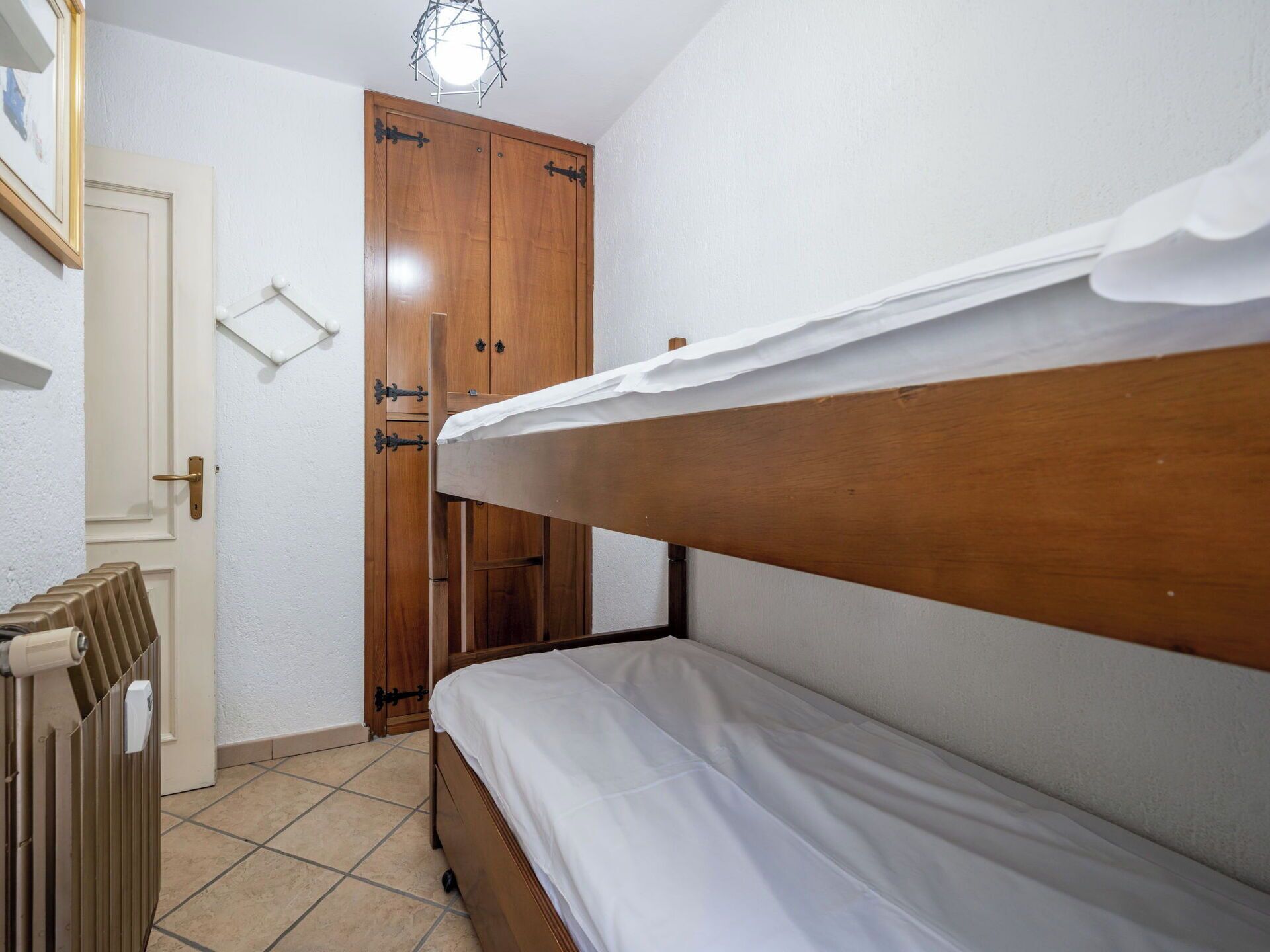 A small, single-bed room with a wooden headboard, a radiator, a wardrobe, and a door with a light fixture above it.