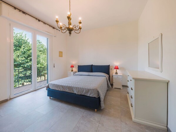 A bedroom with a bed, two bedside tables, a dresser, a chandelier, and a sliding glass door leading to a balcony.