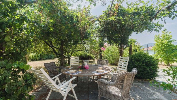 An outdoor dining area with a round table and wicker chairs, surrounded by lush greenery and a view of the sea.