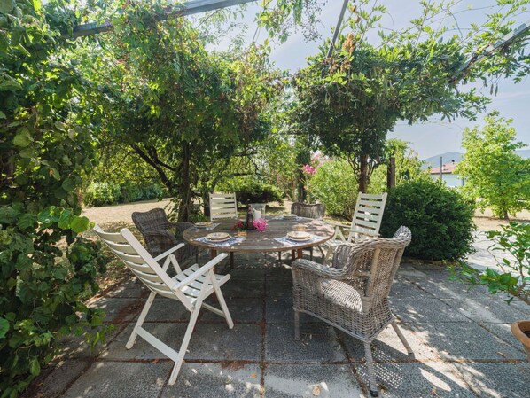 An outdoor dining area with a round table and wicker chairs, surrounded by lush greenery and a view of the sea.