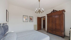 A bedroom with a large bed, a wooden wardrobe, a chandelier, and two framed artworks on the wall.