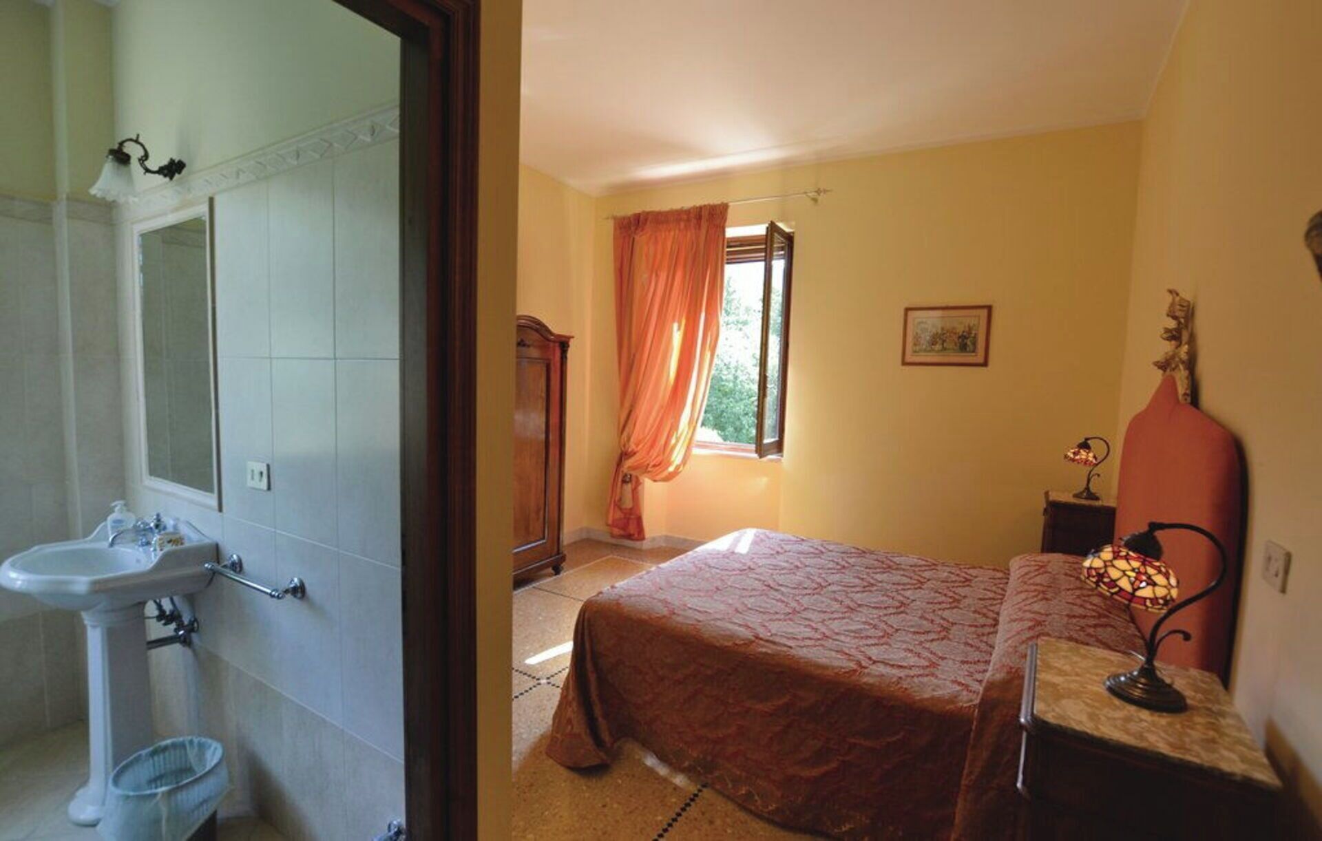 A bathroom with a sink and mirror, a bedroom with a bed and window, and a nightstand with a lamp.