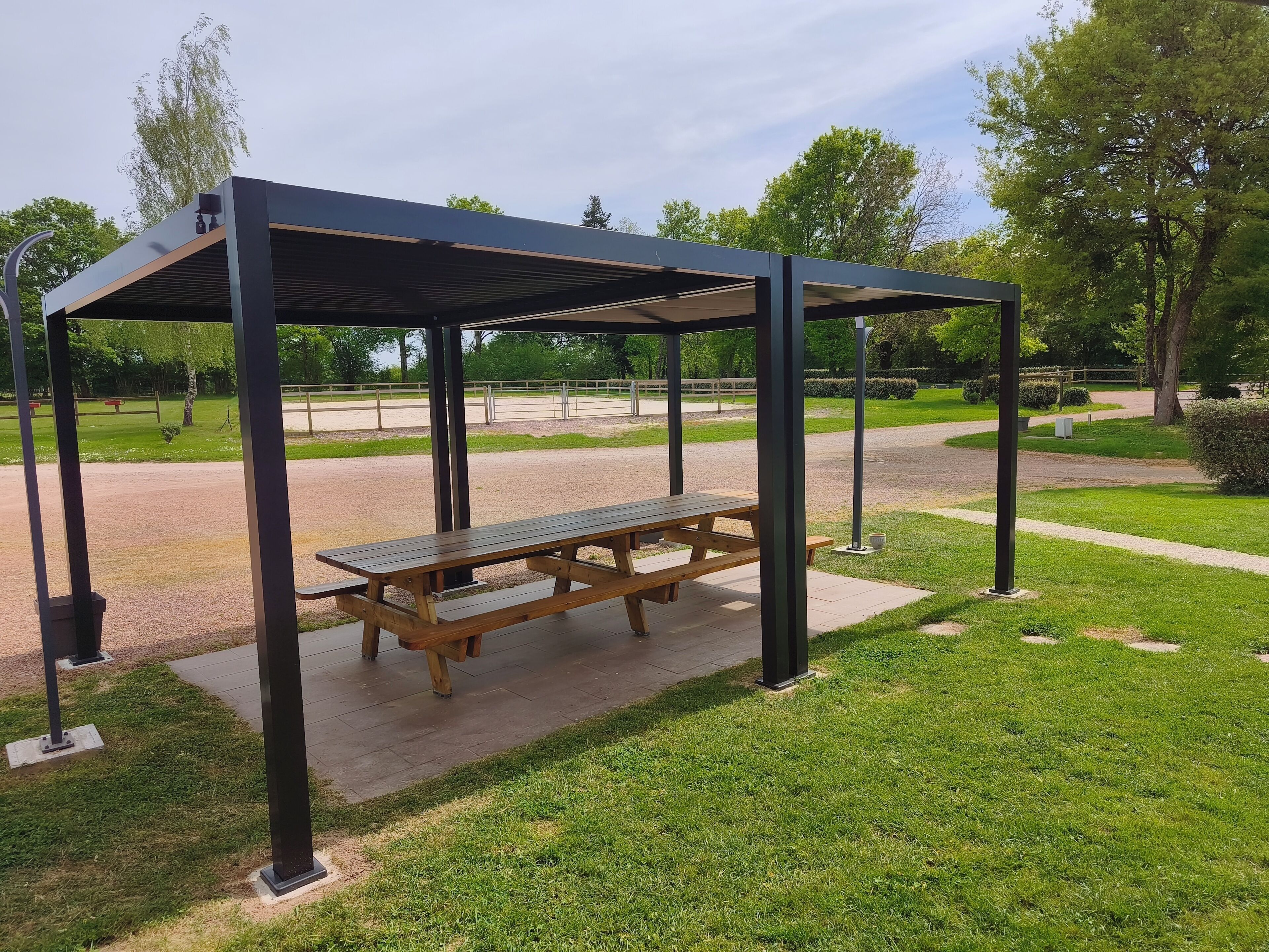 Un área de picnic techada con un banco, rodeada por un césped cuidado y un campo deportivo cercado.