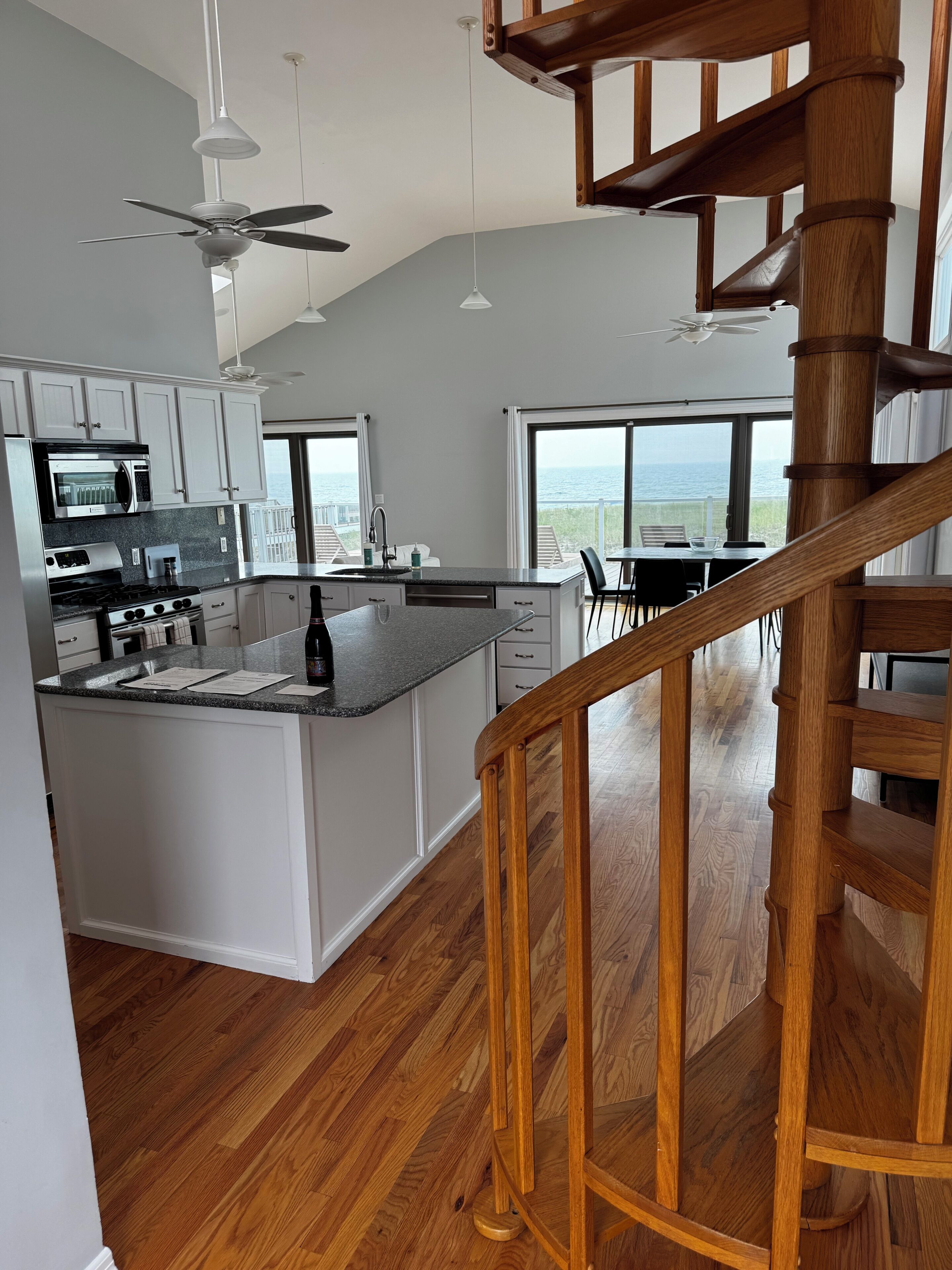 A modern kitchen with a central island, wooden flooring, and a view of the ocean through large windows.