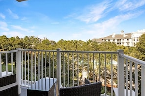 Ein Balkon mit Korbmöbeln und einer weißen Brüstung, der auf eine angelegte Grünfläche mit Bäumen und Gebäuden blickt.