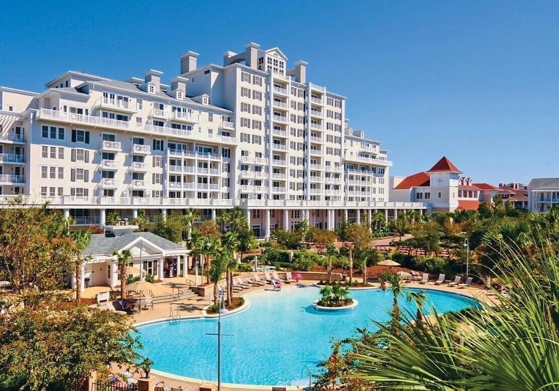 A hotel with a large swimming pool surrounded by lounge chairs and palm trees.