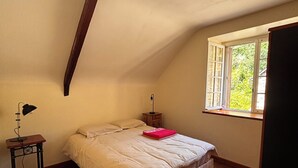 A bedroom with a sloped ceiling, a bed with a beige comforter, a nightstand with a lamp, and a window with a view of greenery.