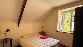 Een slaapkamer met een schuine kap, een bed met een beige dekbed, een nachtkastje met een lamp en een raam met uitzicht op groen.