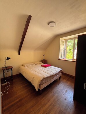 A bedroom with a sloped ceiling, a bed with a beige comforter, a nightstand with a lamp, and a window with a view of greenery.