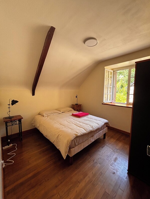 A bedroom with a sloped ceiling, a bed with a beige comforter, a nightstand with a lamp, and a window with a view of greenery. - 600 meters from the beach, a former farmhouse that sleeps 5; pets allowed. (Erquy)