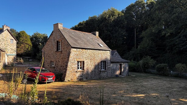 A stone house with a red car parked in front, surrounded by a grassy yard and trees.