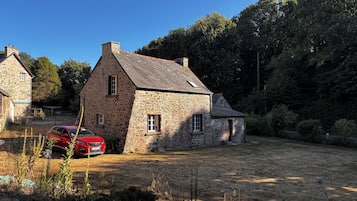 Een stenen huis met een rode auto ervoor, omringd door een gazon en bomen.