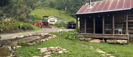 Una cabaña rústica con porche, rodeada de un jardín y un tractor al fondo.