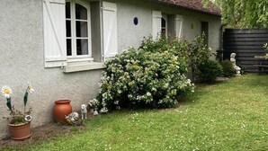 Ett hus med ett tak täckt av tegelpannor, vita fönsterluckor och en trädgård med blommor och en liten staty.