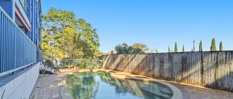 Une piscine à bordures courbes, entourée d’une terrasse en briques et d’une clôture en bois.