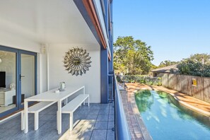 Un espace aménagé au bord de la piscine, avec une table et des bancs blancs, un élément décoratif en métal fixé au mur, et un ciel d’un bleu limpide.