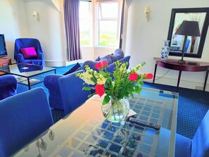 A hotel room with a glass coffee table, blue sofas, a TV, a vase with flowers, and a lamp. - Apartment Sleeps 4 With Garden (Prestwick)