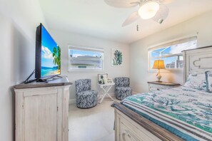 Ein Schlafzimmer mit einem Bett, einem Fernseher, einem Deckenventilator und einem Fenster mit Blick auf Gebäude.