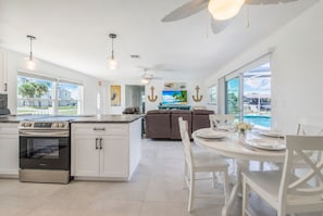 Una cocina moderna con una isla central, comedor con sillas blancas y vista a una piscina.