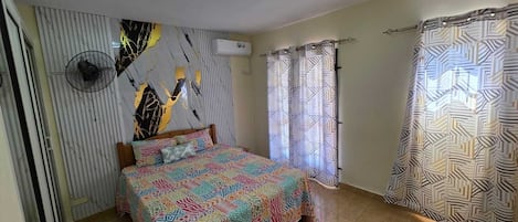 Une chambre à coucher comprenant un lit, un climatiseur fixé au mur, un luminaire au plafond et une fenêtre avec des rideaux.