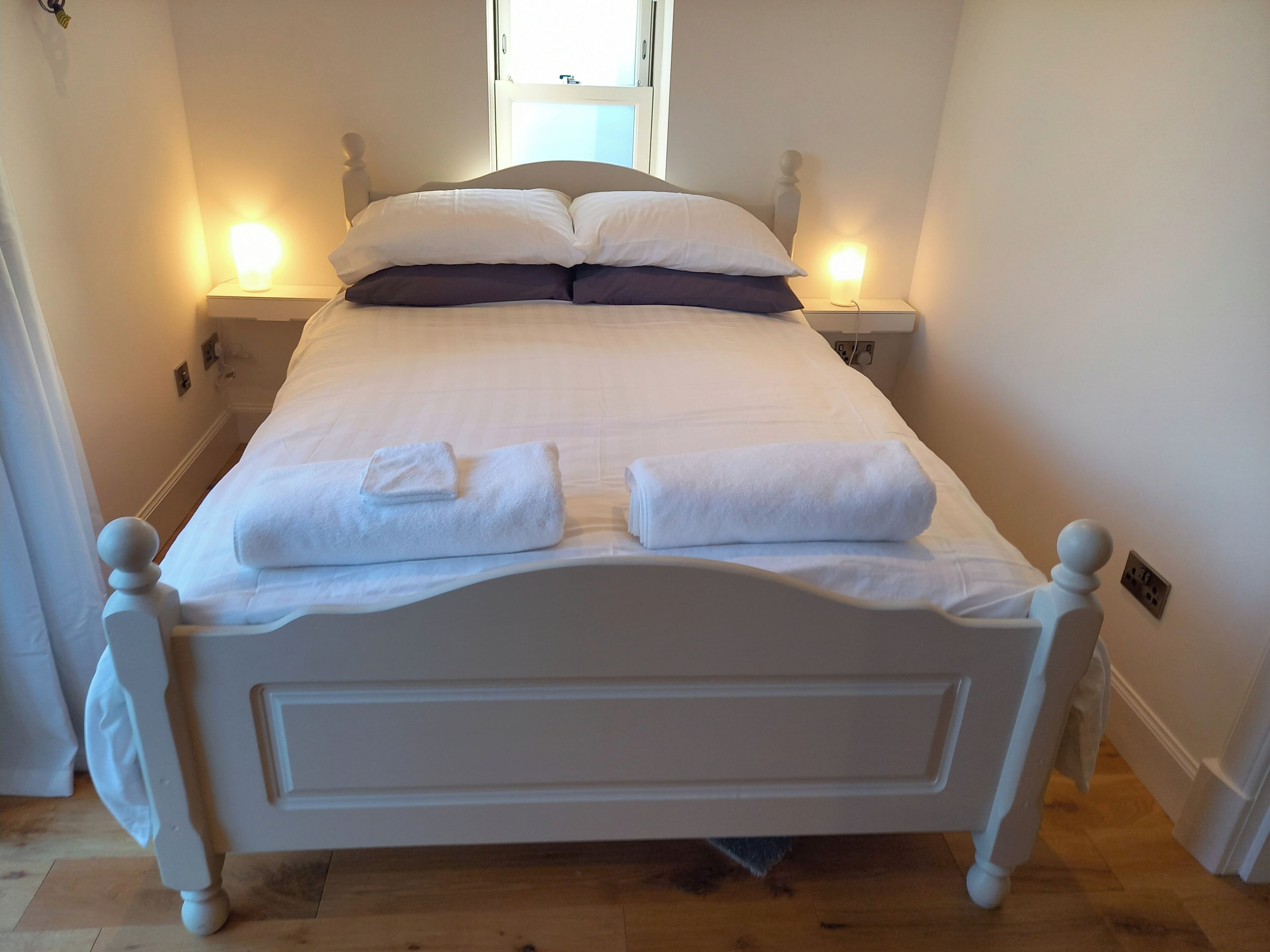 A neatly made bed with white linens and two pillows, a bedside table with a lamp, and a window above the bed.