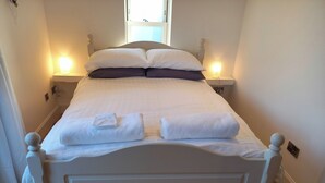 A neatly made bed with white linens and two pillows, a bedside table with a lamp, and a window above the bed. - Holiday Cottage Sleeps 3 With Garden (Prestwick)